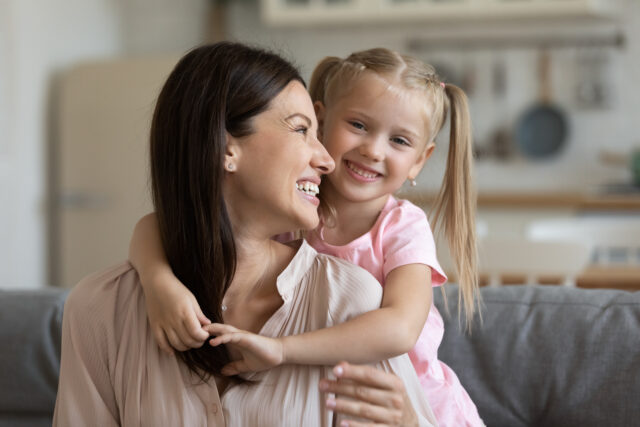 Happy small blonde preschool girl cuddling from back smiling young mother, looking at camera. Head shot portrait bonding family of two enjoying spending leisure weekend time together at home.